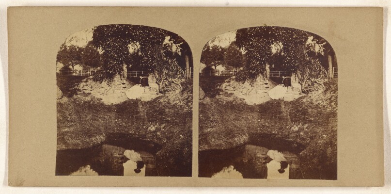 [Woman seated on ground overlooking a brook, her reflection seen in the brook]