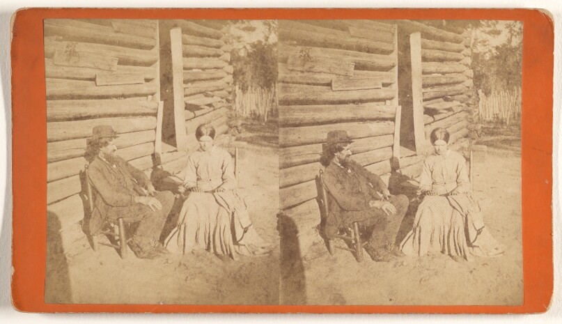 [Man and woman seated in front of log cabin]