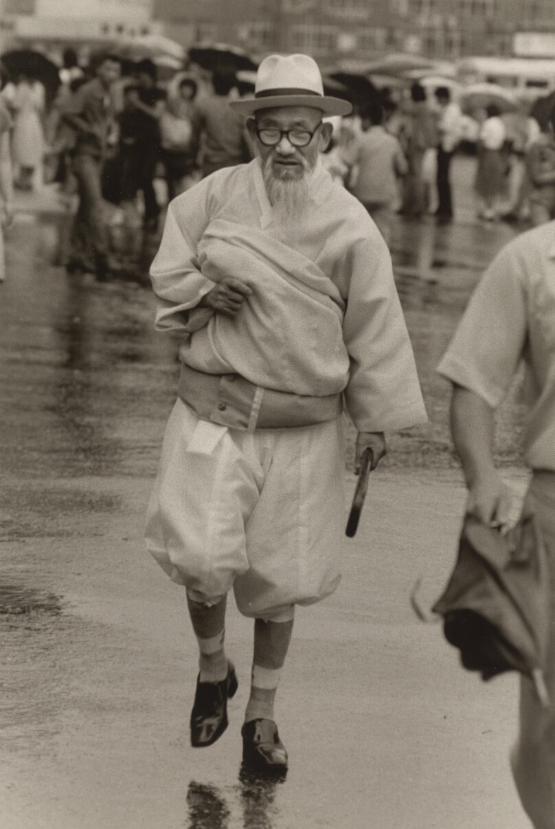 [White-Bearded Man in Glasses and Straw Hat and White Traditional Garb Holding His Long Robe Up While Walking Through the Rain]