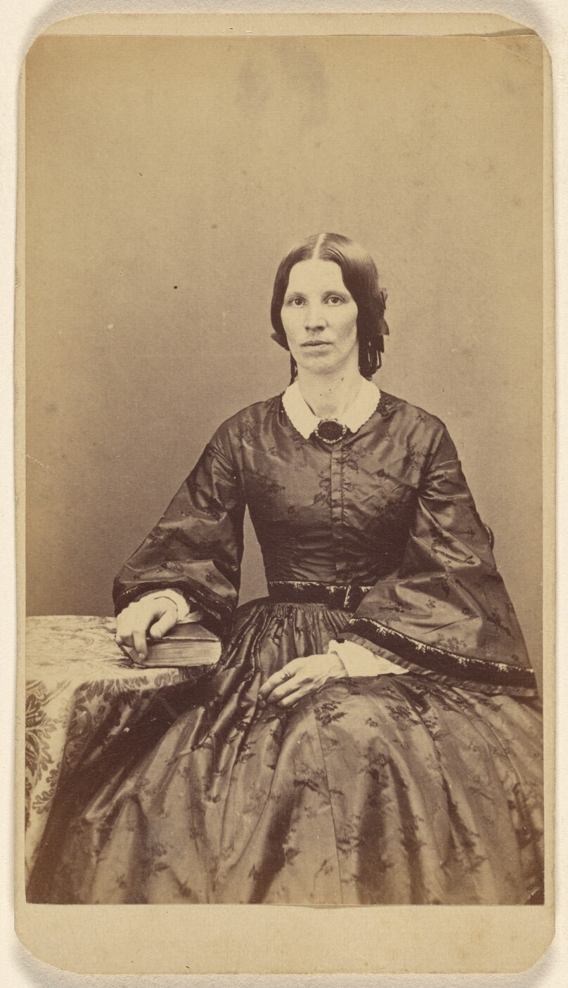 [Unidentified woman seated, resting a hand on a book on a table]