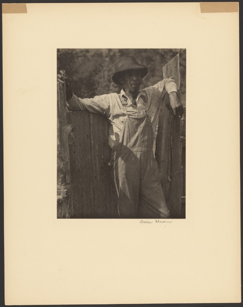 [Man in Overalls Leaning Against Old Fence]