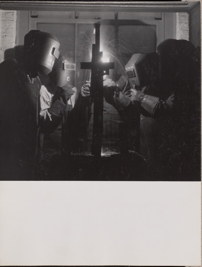 [Four men wearing welding helmets weld a piece of metal]