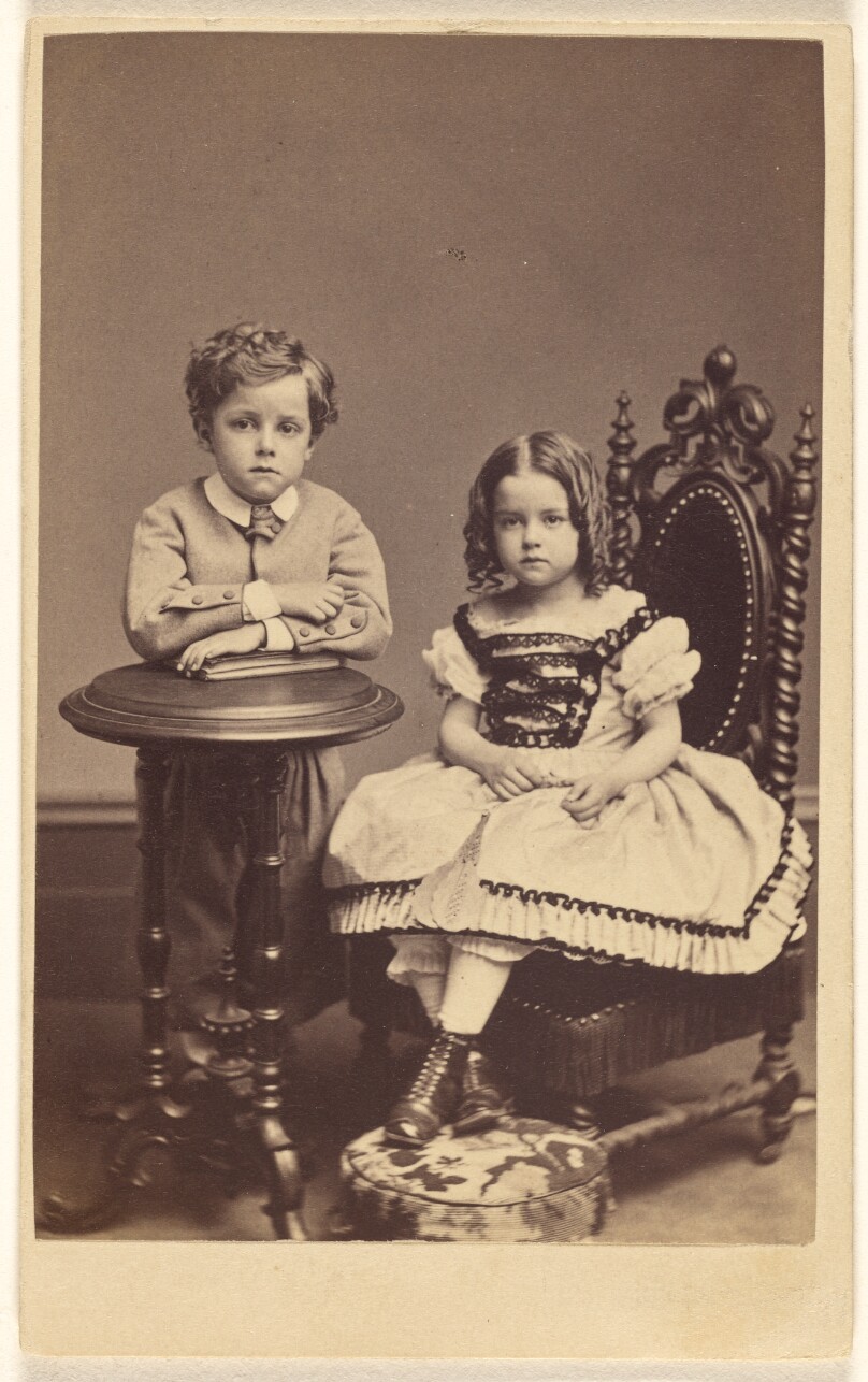 [Two children: boy standing, leaning on a table; girl with curls, seated]