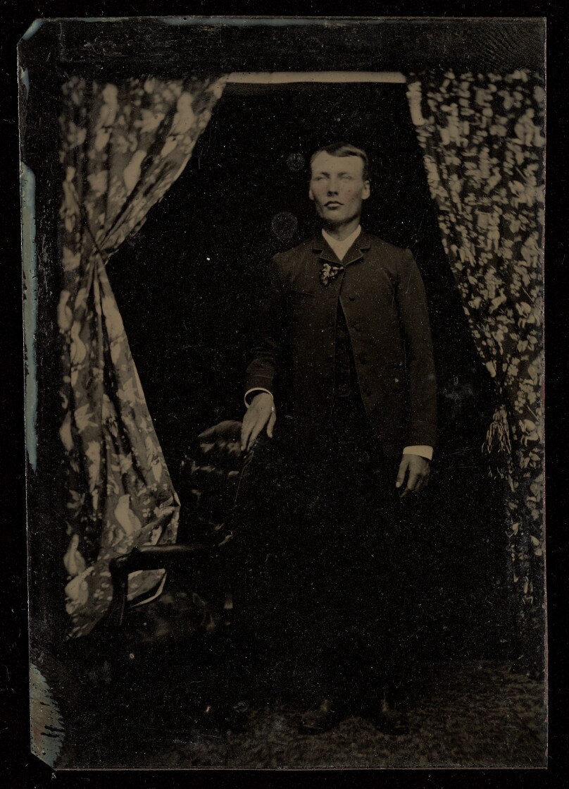 [Unidentified man standing, with one hand on a chair back]
