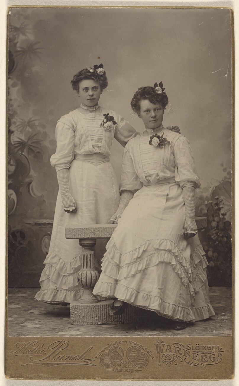 [Two unidentified women wearing identical white dresses, both with flowers in hair and on dresses]