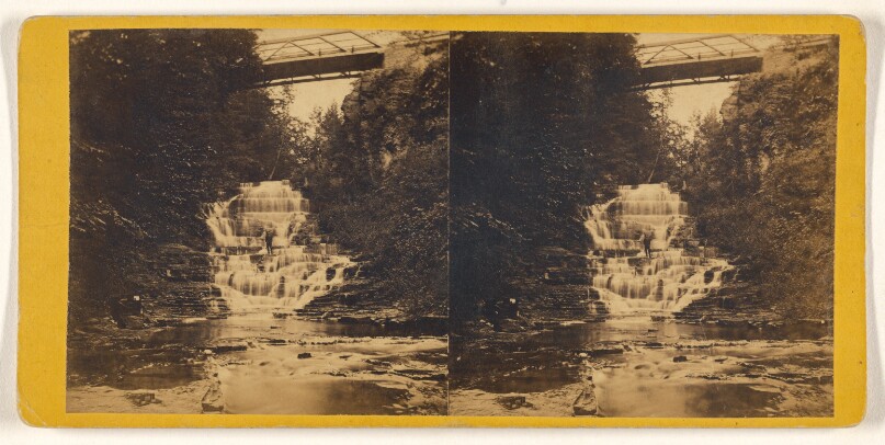 [Unidentified falls with bridge in background, man standing in center of stream]