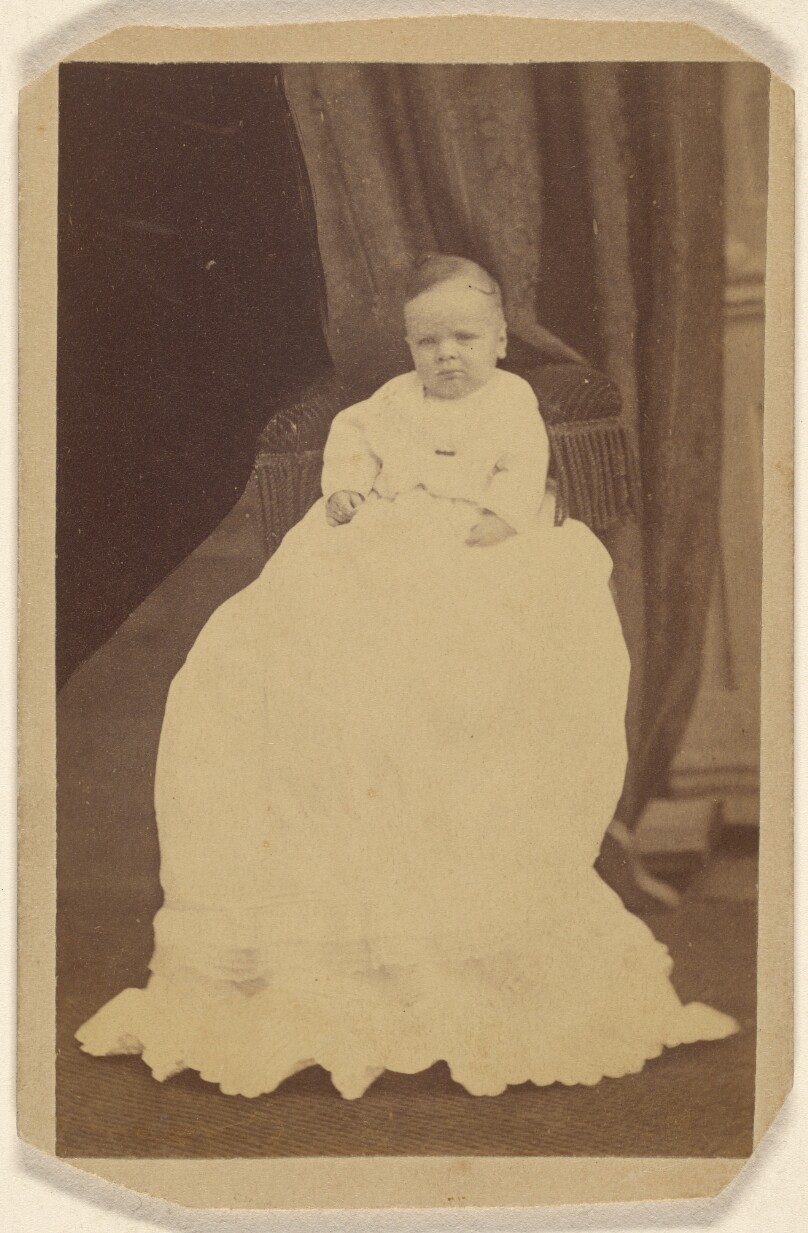 [Unidentified baby wearing a long gown, seated on a couch]
