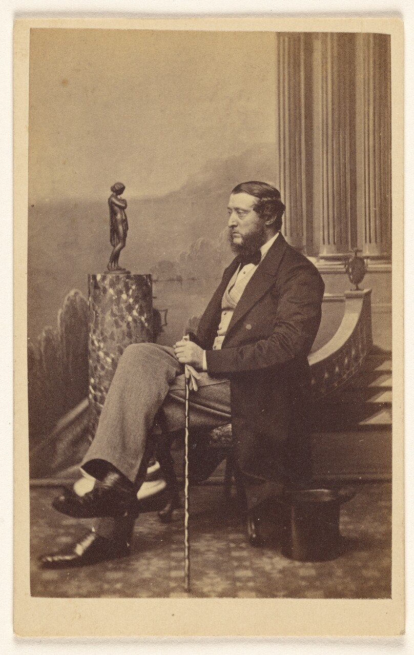 [Unidentified bearded man seated, holding a walking stick]