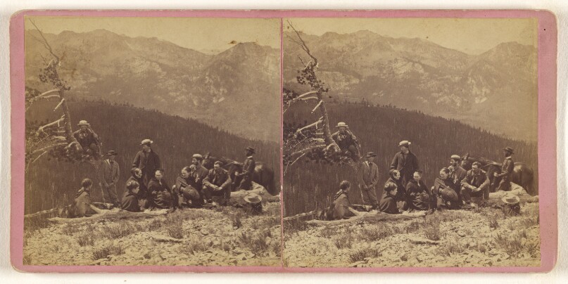 [Group of people on a hillside, mountains and valleys in background, one man is in a tree, another man standing by a horse]