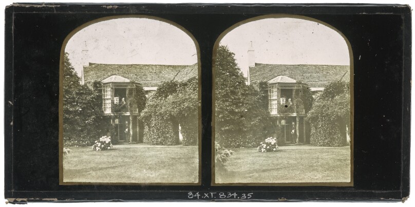 [View of a two-storey British cottage and garden with a man standing at the ground doorway and a woman seated on the second level balustrade]